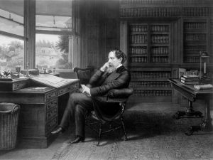 A black and white portrait of Charles Dickens pondering at his writing desk in his study.